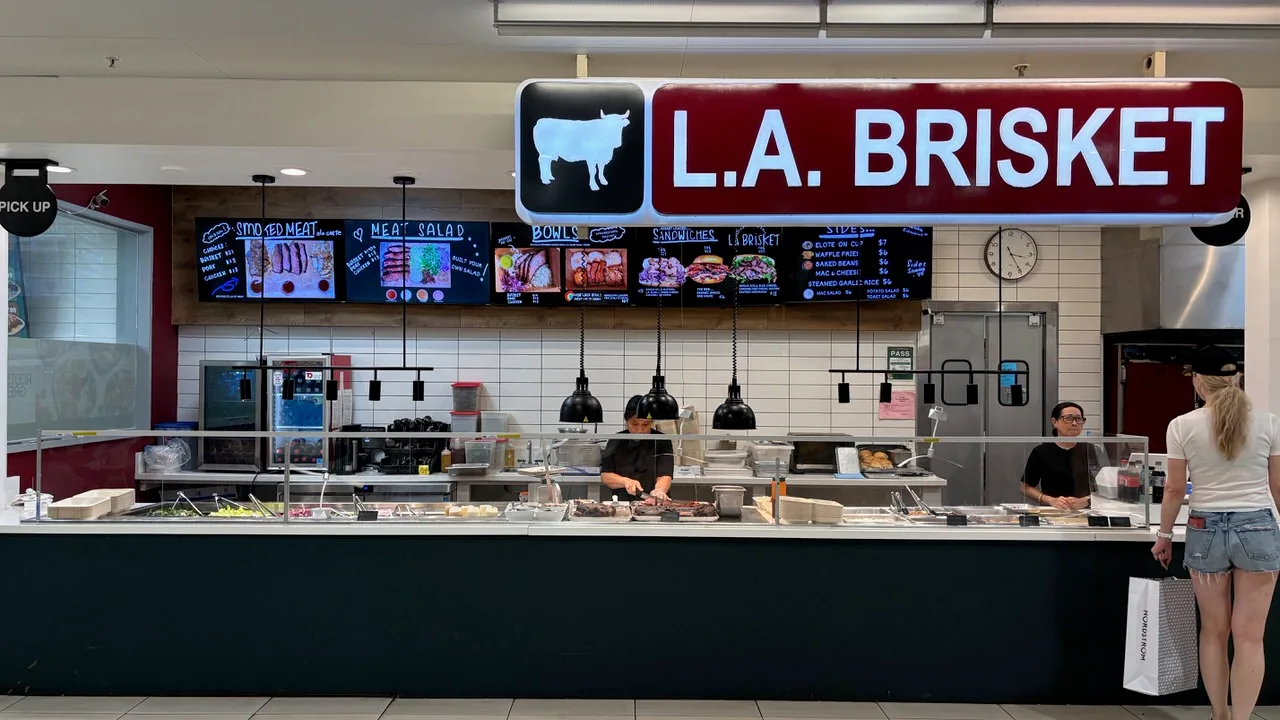 The L.A. Brisket counter under a red sign with a white cow, menu screens for smoked meat, meat salad, bowls, sandwiches and sides above a glass-fronted serving line