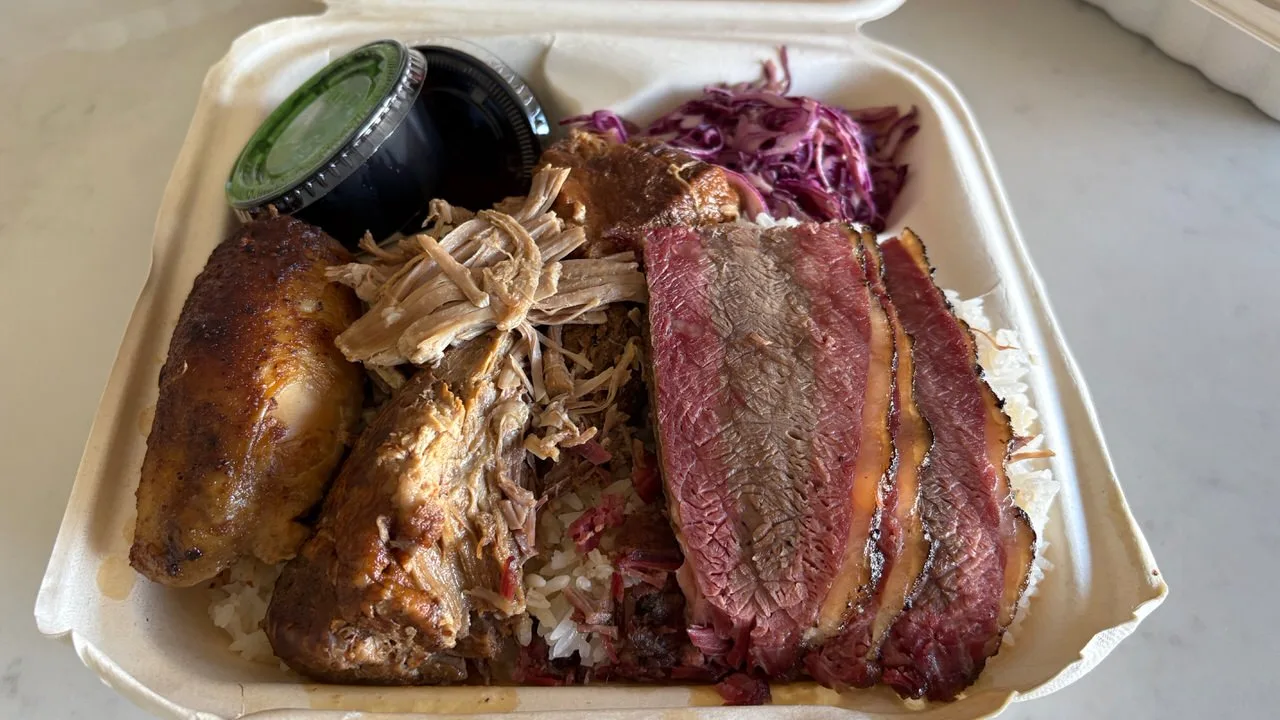 Sliced smoked brisket, pulled pork and roasted chicken over rice in a takeout box, with purple cabbage slaw and two lidded sauce cups