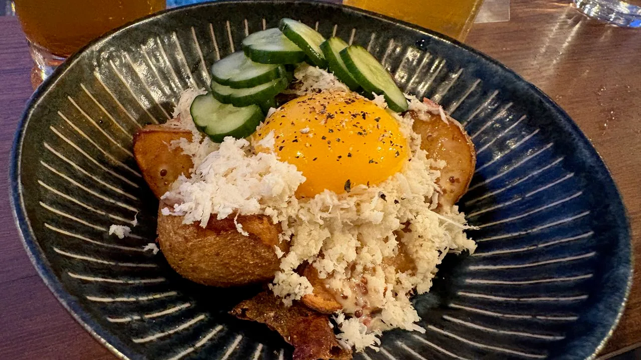 A dark blue ridged bowl of fried potato pieces topped with grated white horseradish, a raw egg yolk with black pepper, and sliced cucumber pickle