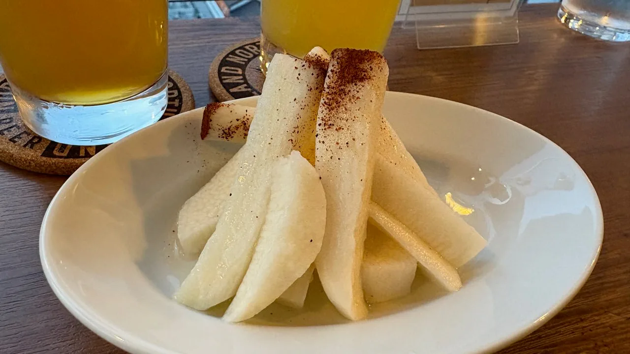 Pale batons and slices of nagaimo yam piled in a shallow white bowl, dusted with a dark reddish powder, with two glasses of beer behind