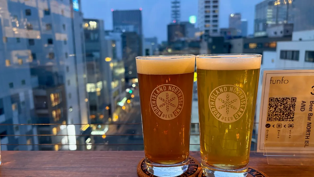 Two glasses of beer on a window counter, one amber and one pale gold, with lit city buildings behind them at dusk