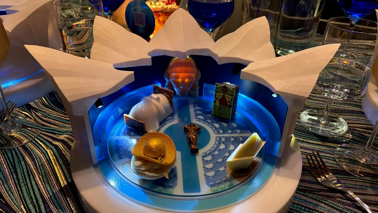 A white model of the LUDÕ theater opened out, its blue-lit circular base holding four small desserts around a bronze figure