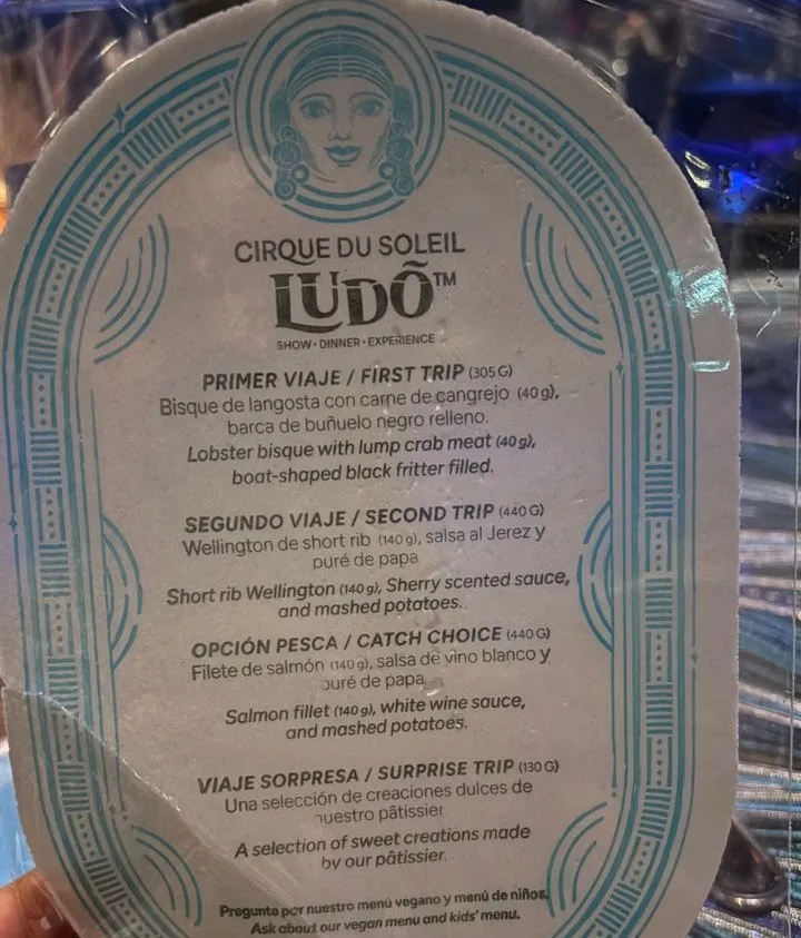An oval menu card printed in Spanish and English with a blue line-drawn face at the top, listing four LUDÕ courses with gram weights