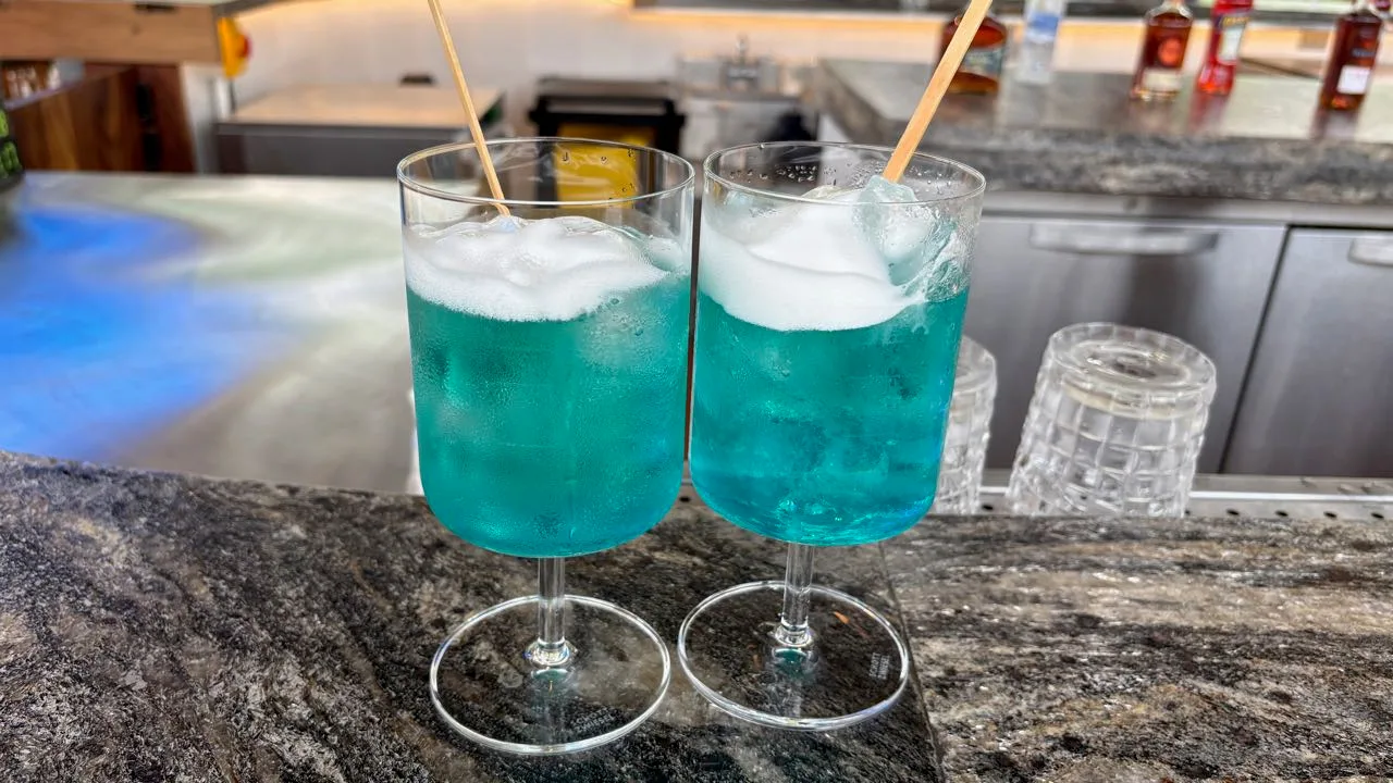 Two stemmed glasses of turquoise drink with a white foam layer and wooden straws, on a stone bar counter