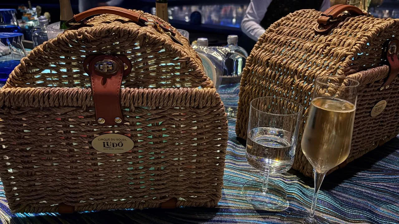 Two wicker picnic baskets with leather straps and a small Cirque du Soleil LUDÕ badge, beside a glass of water and a flute of champagne