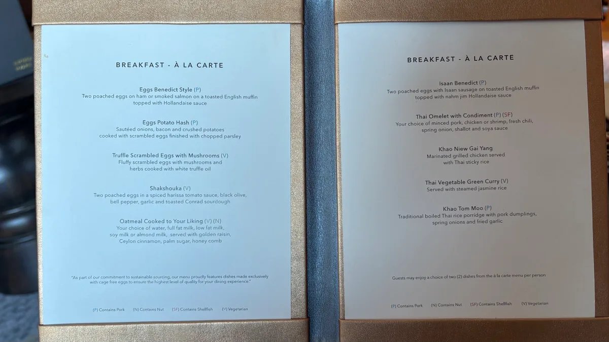 An open menu with two pages headed Breakfast - À la carte: eggs Benedict, potato hash, truffle scrambled eggs, shakshouka and oatmeal on the left; Isaan Benedict, Thai omelet, Khao Niew Gai Yang, Thai vegetable green curry and Khao Tom Moo on the right