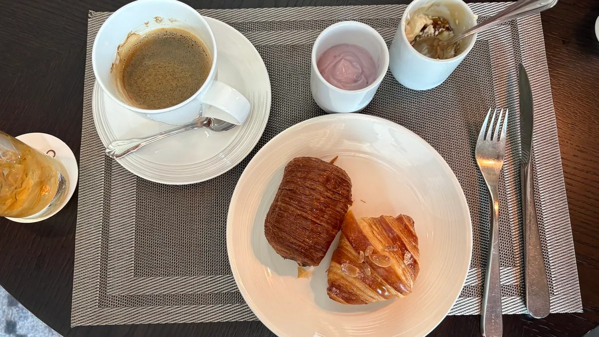 A breakfast table from above: a cup of coffee, a plate with an almond croissant and a dark pastry, a cup of purple yogurt, and at the top right a cup of yogurt with honeycomb and a spoon