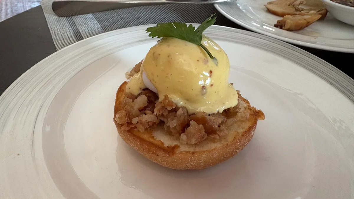 Half an English muffin topped with crumbled sausage meat, a poached egg under hollandaise sauce and a coriander leaf, on a white plate