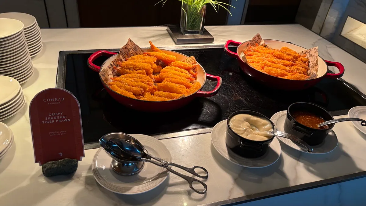 Two red pans of breaded fried prawns on a buffet counter, a Conrad Bangkok sign reading Crispy Shanghai Tiger Prawn, and pots of a white dip and a sweet chili sauce