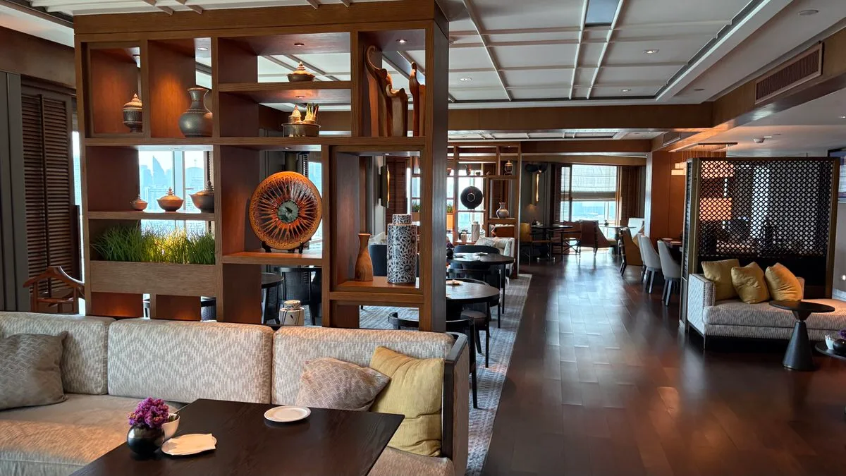A hotel lounge with dark wood shelving holding vases and ceramics, grey sofas with yellow cushions, dining tables and tall windows onto city buildings