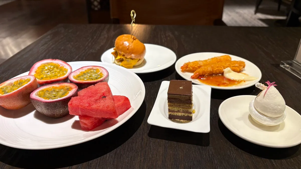 Plates on a dark table: halved passion fruit and watermelon, a slider on a skewer, two fried prawns with sauces, a layered chocolate cake and a small white dessert