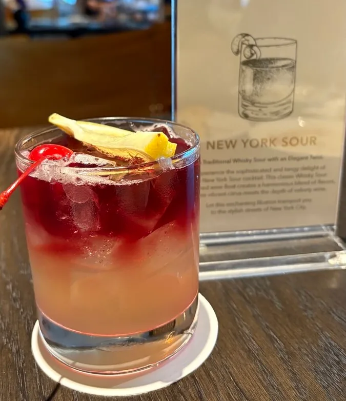 A cocktail in a short glass, deep red on top and paler below, with ice, a lemon peel and a cherry, in front of a card reading New York Sour