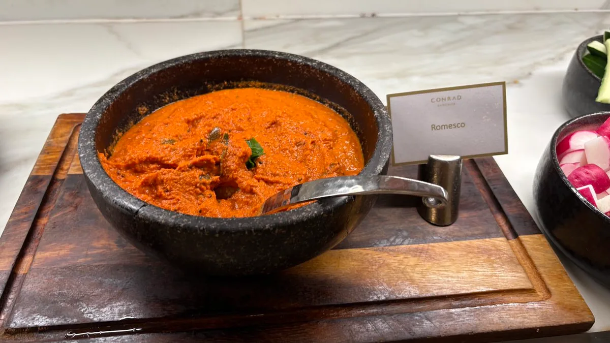 A dark stone bowl of thick orange-red sauce with a ladle on a wooden board, beside a Conrad Bangkok card reading Romesco and a bowl of sliced radishes