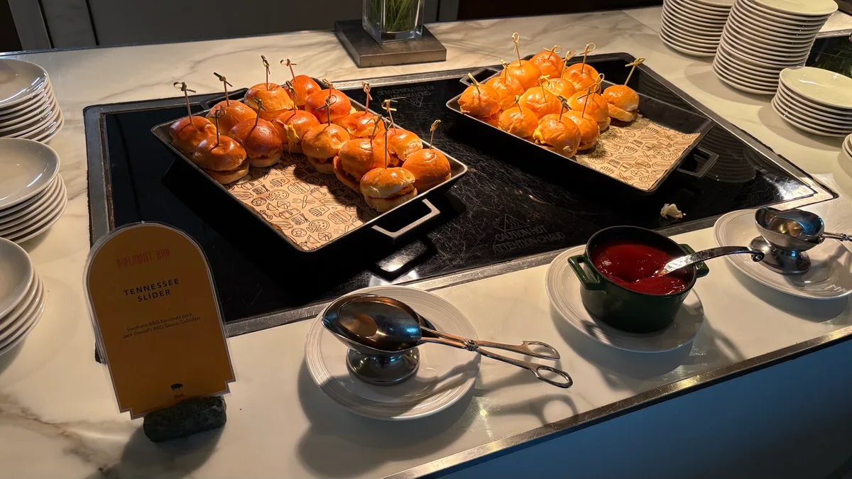 Two trays of small burgers held together with wooden skewers on a buffet counter, a Diplomat Bar sign reading Tennessee Slider, and a pot of red sauce