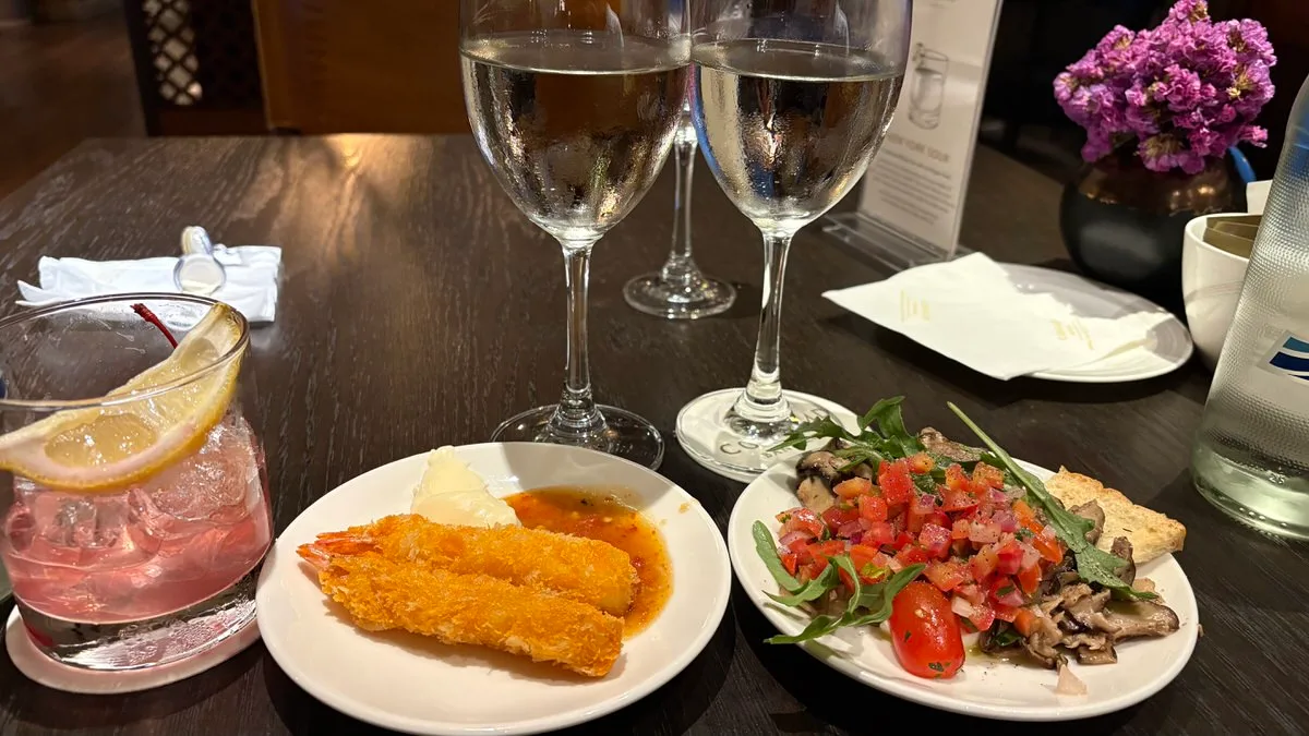 Two glasses of white wine on a dark table, a plate of two fried prawns with sauces, a plate of tomato salsa, mushrooms, rocket and toast, and a pink drink with a lemon slice at the left
