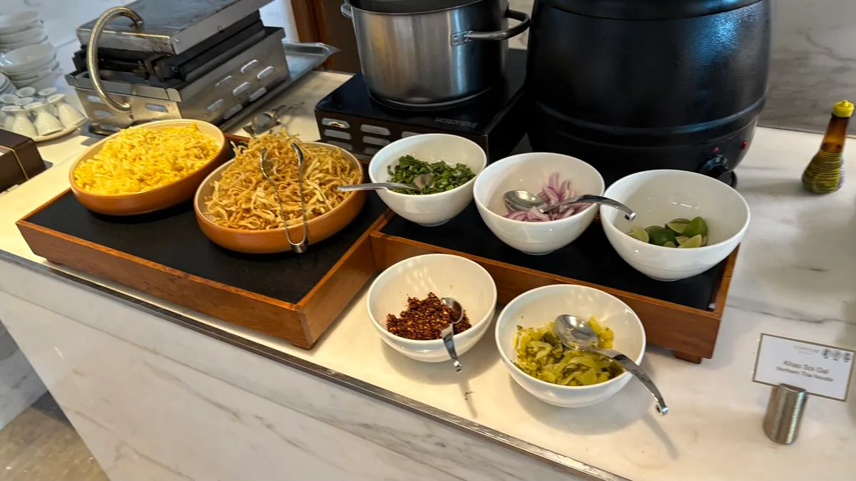A buffet counter with two wooden bowls of noodles, one crispy fried, small white bowls of chopped greens, sliced red onion, lime wedges, chili flakes and pickled mustard greens, soup pots behind and a small name card