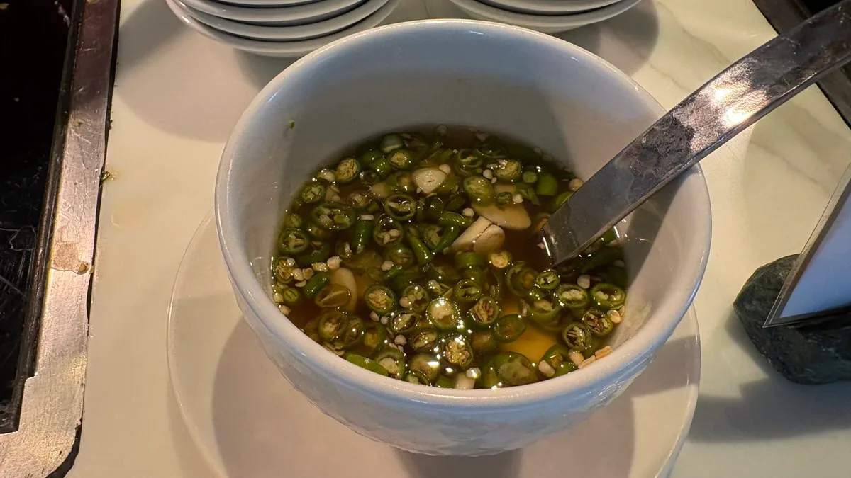 A white bowl of brown liquid full of sliced green chilies and pieces of garlic, with a metal spoon, on a saucer