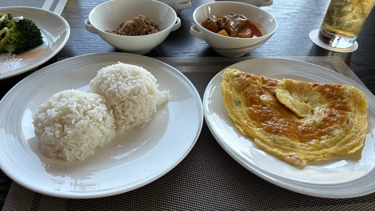 A folded golden omelet on one plate and two mounds of steamed white rice on another, with bowls of minced meat and beef curry behind and a glass of iced tea