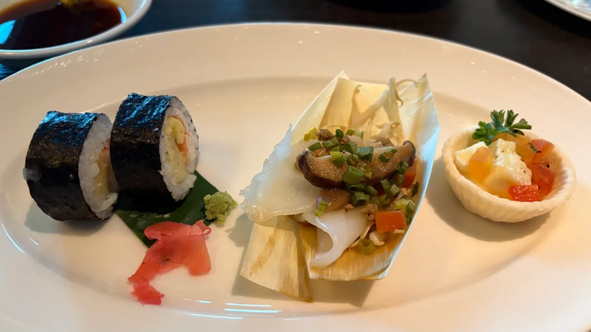 A white plate with two pieces of nori-wrapped sushi roll on a green leaf with wasabi and pink pickled ginger, a dried-leaf boat of white slices topped with mushroom and chopped green onion, and a small pastry cup of diced orange, red and white pieces with parsley; a dish of soy sauce at the top left