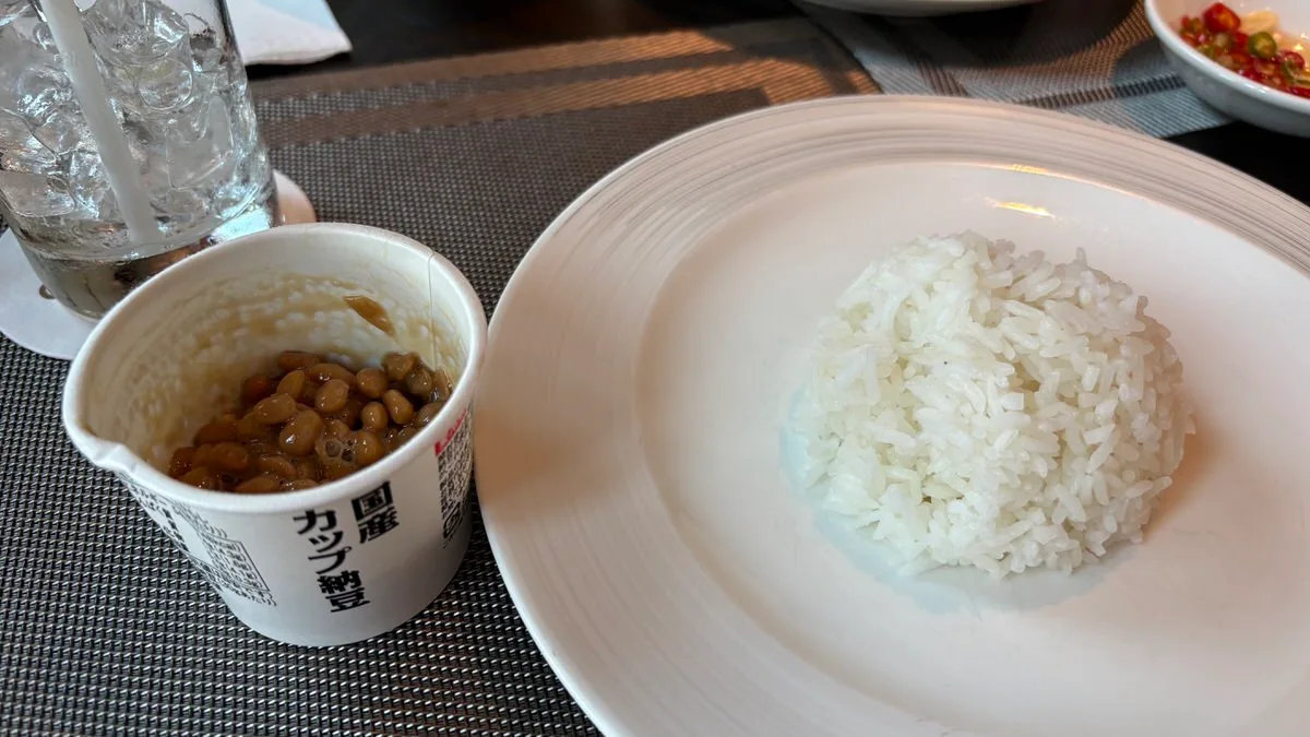 A paper cup of natto with Japanese lettering beside a mound of white rice on a white plate, with a glass full of ice and a small dish of chopped chilies at the edges