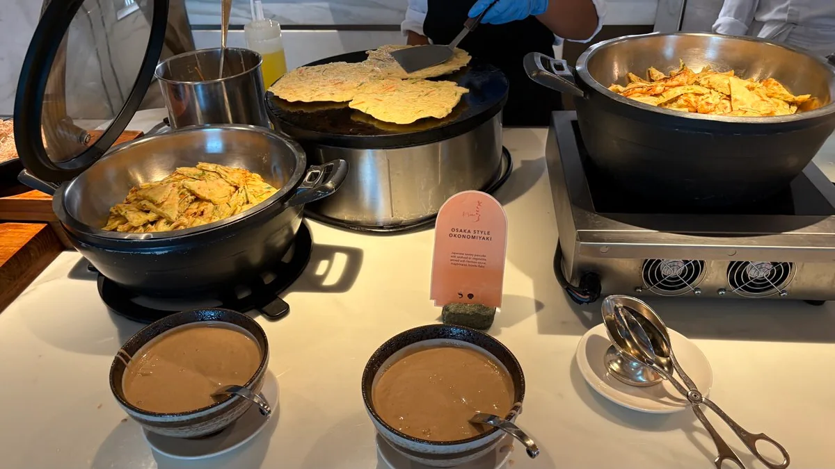 A buffet station with thin pancakes on a round griddle under a spatula held in a blue-gloved hand, two black pans of cut pancake pieces, a card reading Osaka Style Okonomiyaki, and two bowls of light brown sauce in front