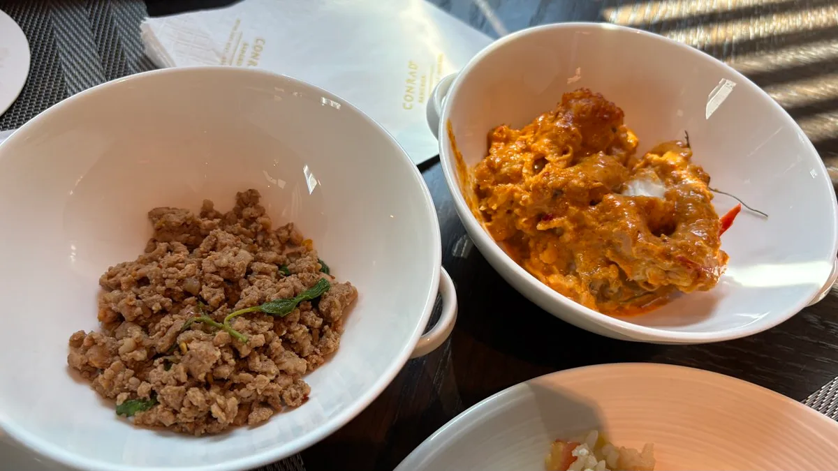 Two white bowls on a dark table: minced pork with a few basil leaves on the left and pieces of fish in a thick orange-red curry on the right, with a Conrad paper napkin behind