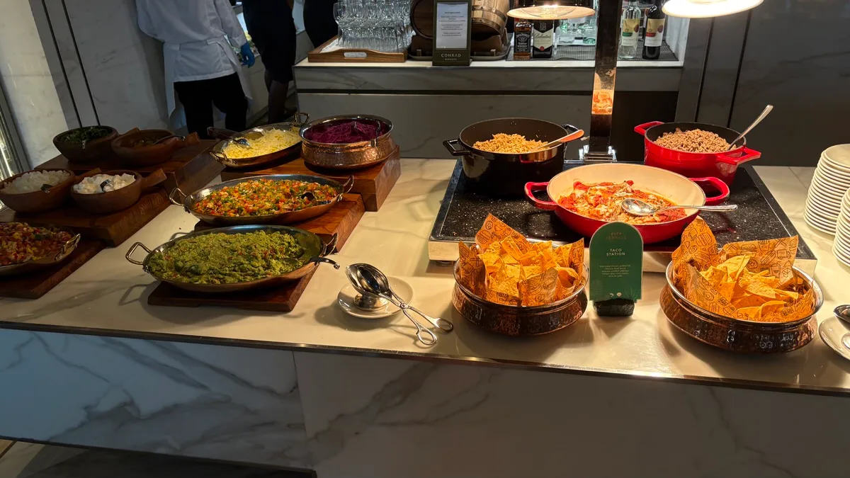 A buffet counter with pans of guacamole, diced salsas, grated cheese and purple cabbage on wooden boards, three pots of fillings on a hot plate, and two copper bowls of tortilla chips