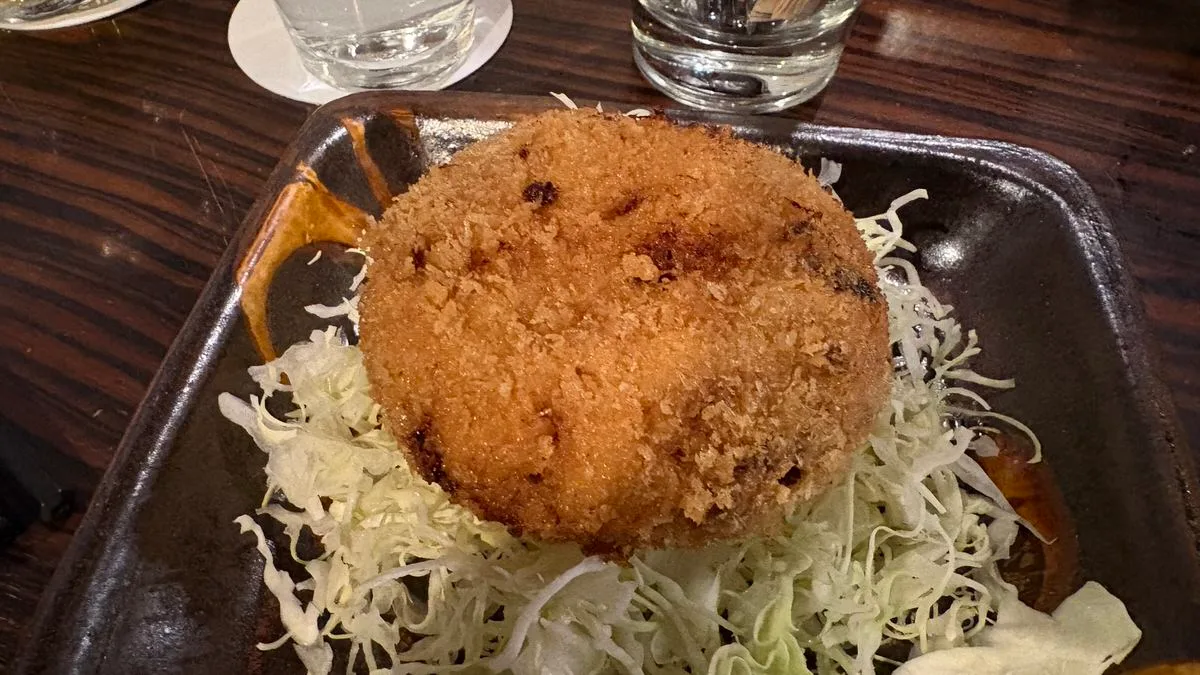 A round crumbed and deep-fried cutlet, golden brown and whole, resting on shredded cabbage in a dark square dish