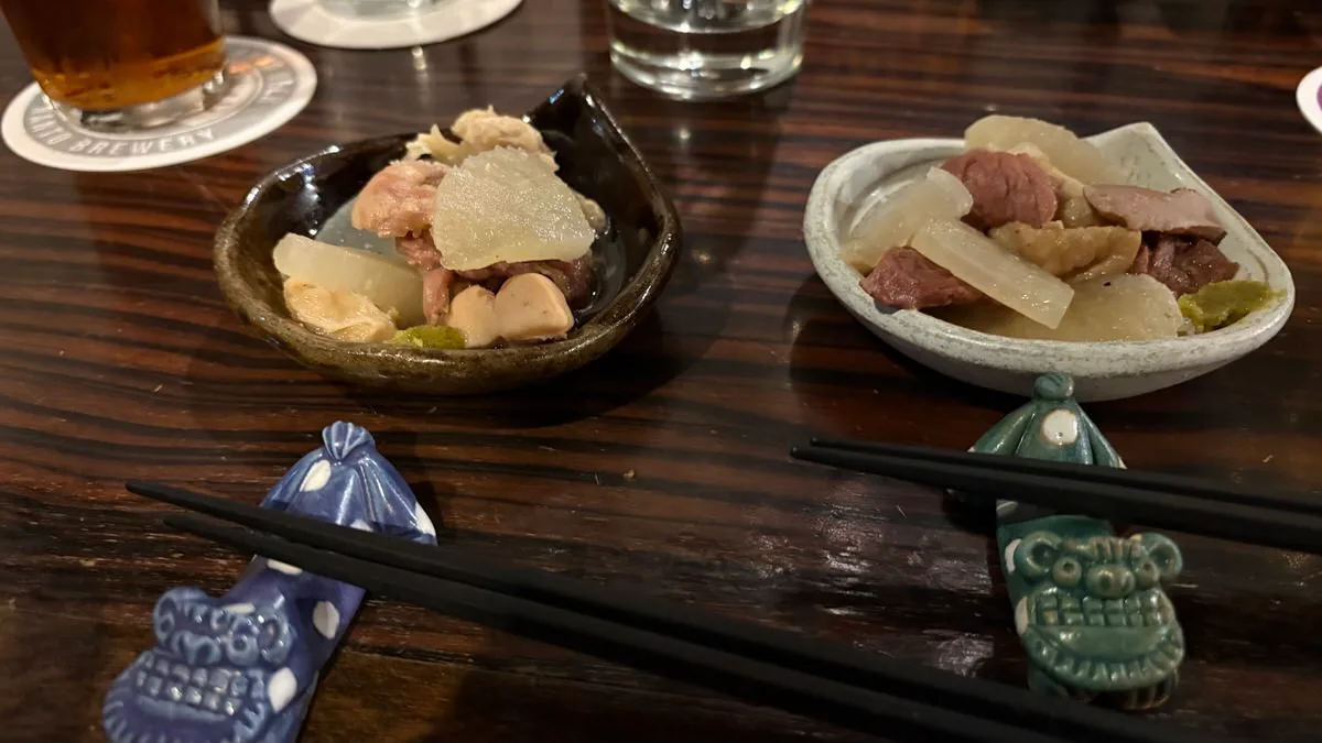 Two small dishes of stewed meat and pale simmered vegetable with a dab of green paste, on a dark wooden counter with chopsticks on shisa-shaped rests