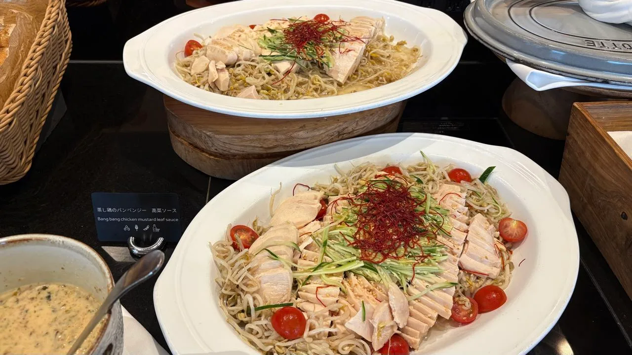Two oval platters of sliced steamed chicken over bean sprouts with cucumber, halved cherry tomatoes and red chilli threads, with a bowl of pale sauce beside them