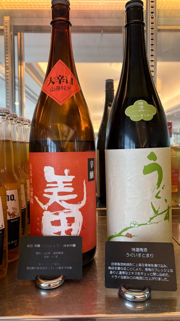 Two large bottles on a shelf, a red-labelled sake with a neck tag and a white-labelled plum wine, each with a Japanese description card, more bottles to the left