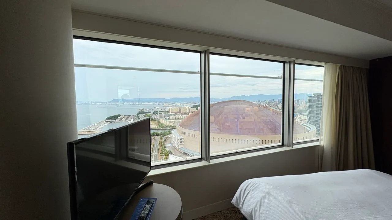 The roof of a large domed stadium seen through a hotel room window, with the bay to the left and a bed in the foreground