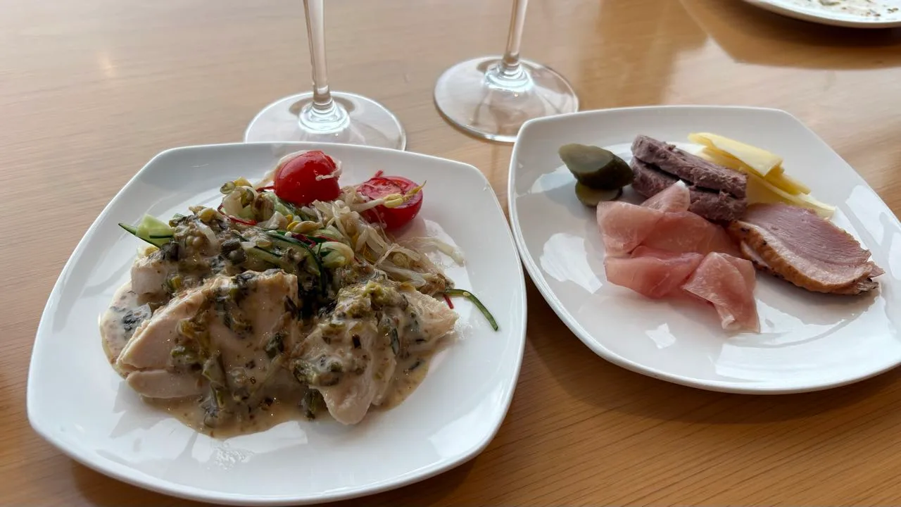 Two white plates on a wooden table, one with bang bang chicken under a pale green sauce, one with prosciutto, sliced duck, salami, cheese and a pickle, wine glasses behind