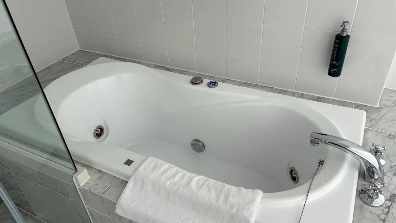 A white bathtub with jet nozzles set into marble tiling, a folded white towel over the rim and a bottle of shower gel on the wall