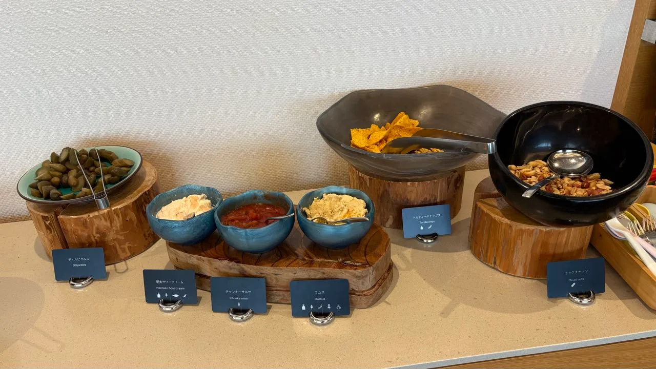 A buffet counter with a bowl of dill pickles, three blue bowls of dips, a bowl of tortilla chips and a black bowl of mixed nuts, each with a small label card