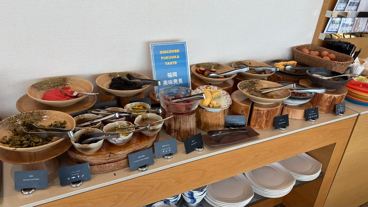 A hotel buffet counter with wooden bowls on log stands, a blue sign reading Discover Fukuoka Taste, a basket of eggs at the far end and labelled cards along the front edge