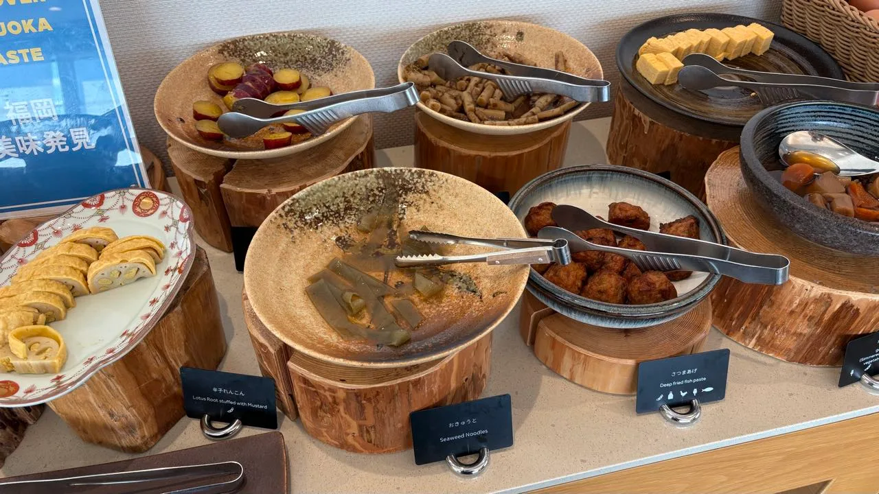 Buffet bowls on wooden log stands holding sliced lotus root, burdock, tamagoyaki, translucent slabs of okyuto and browned fish cakes, with cards reading Lotus Root stuffed with Mustard, Seaweed Noodles and Deep fried fish paste