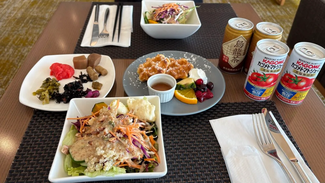 A hotel table with two square bowls of salad, a plate of Japanese pickles, a grey plate with waffles and fruit, and cans of oolong tea and Kagome tomato juice