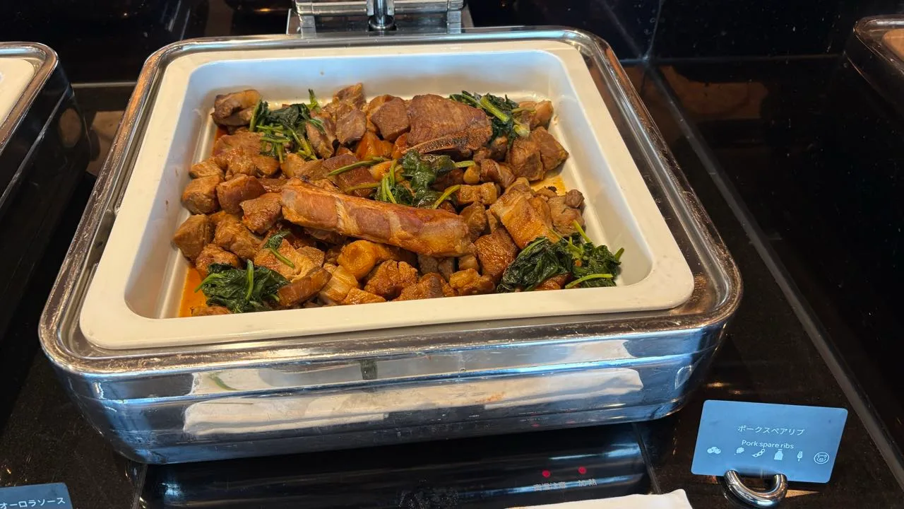 A chafing dish holding chunks of braised pork and one long rib in a dark sauce with green leaves through it, a card in front reading Pork spare ribs