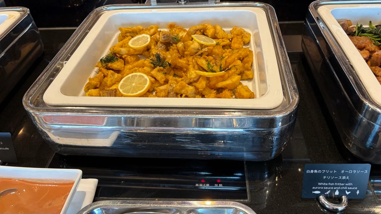 A chafing dish of golden battered pieces of white fish topped with lemon slices and sprigs of dill, with a card reading White fish fritter with aurora sauce and chili sauce