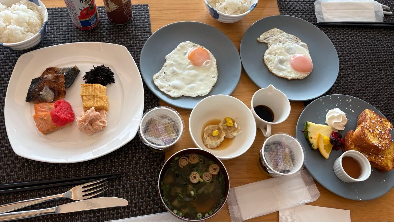 A breakfast table with two bowls of rice, two fried eggs on grey plates, a plate of grilled fish, hijiki, sliced tamagoyaki, salmon topped with pink cod roe, a small bowl of shumai, miso soup and French toast with fruit