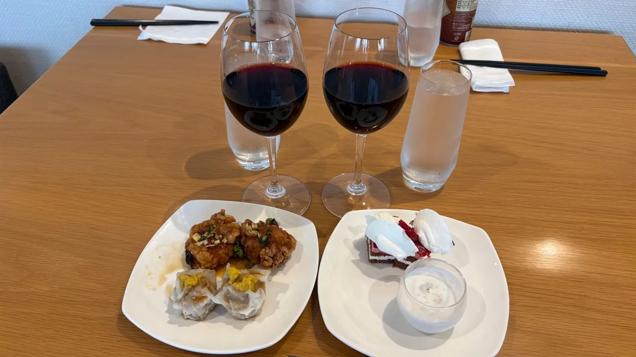 Two glasses of red wine and two water glasses on a wooden table, one plate holding fried chicken and two shumai, another with a slice of chocolate cherry cake and a small glass of white coconut dessert