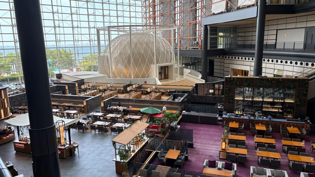 A hotel atrium seen from a fifth-floor walkway: a white latticed dome, restaurant tables and a bar below, and a tall glass wall with the sea beyond