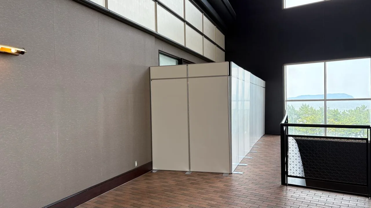 Plain white partition panels closing off a corner of a hotel corridor, with pine trees and the sea visible through the window on the right