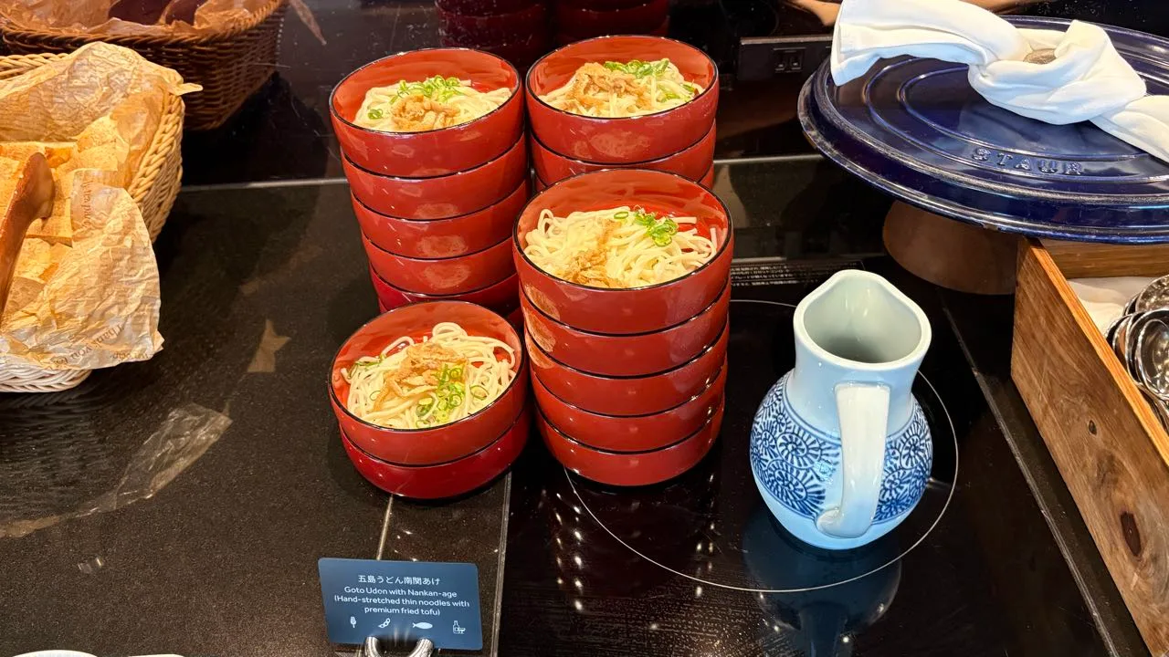Stacks of small red lacquer bowls of cold udon topped with chopped green onion and fried tofu, a blue and white pitcher of broth beside them, and a card reading Goto Udon with Nankan-age