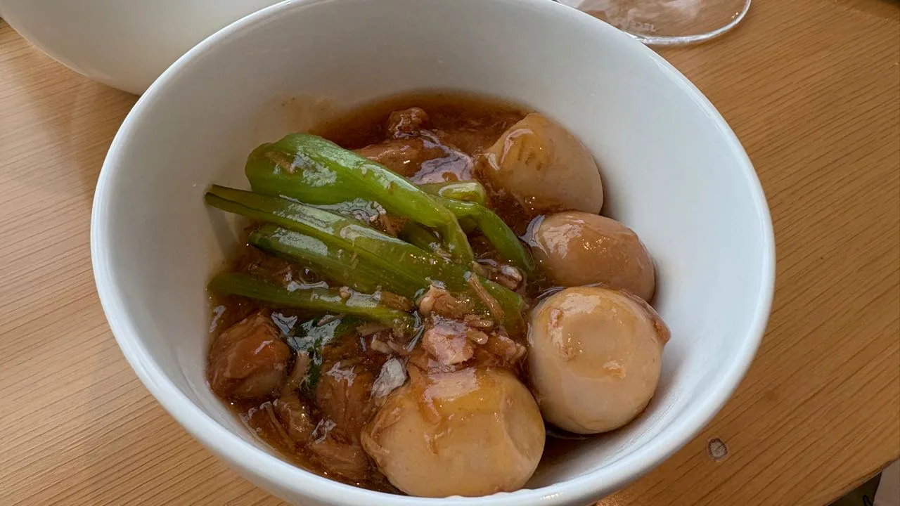 A white bowl of simmered pork in dark soy broth with green stalks on top and four pale quail eggs around the edge