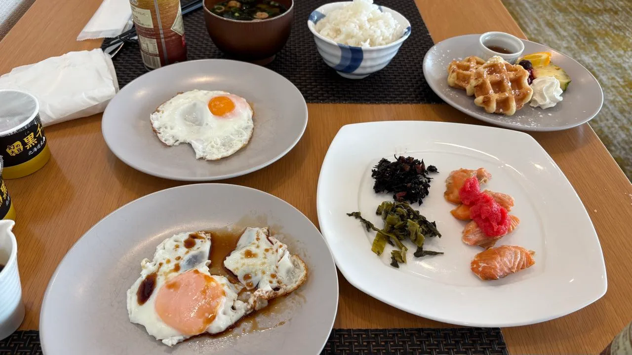 A hotel lounge breakfast table for two: two plates of fried eggs, a bowl of white rice, miso soup, a plate of hijiki, pickled greens and salmon topped with pink cod roe, and a waffle with fruit and whipped cream