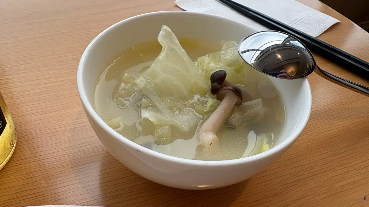 A white bowl of pale broth with cabbage and one shimeji mushroom in it, a metal ladle resting on the rim and black chopsticks on the table behind
