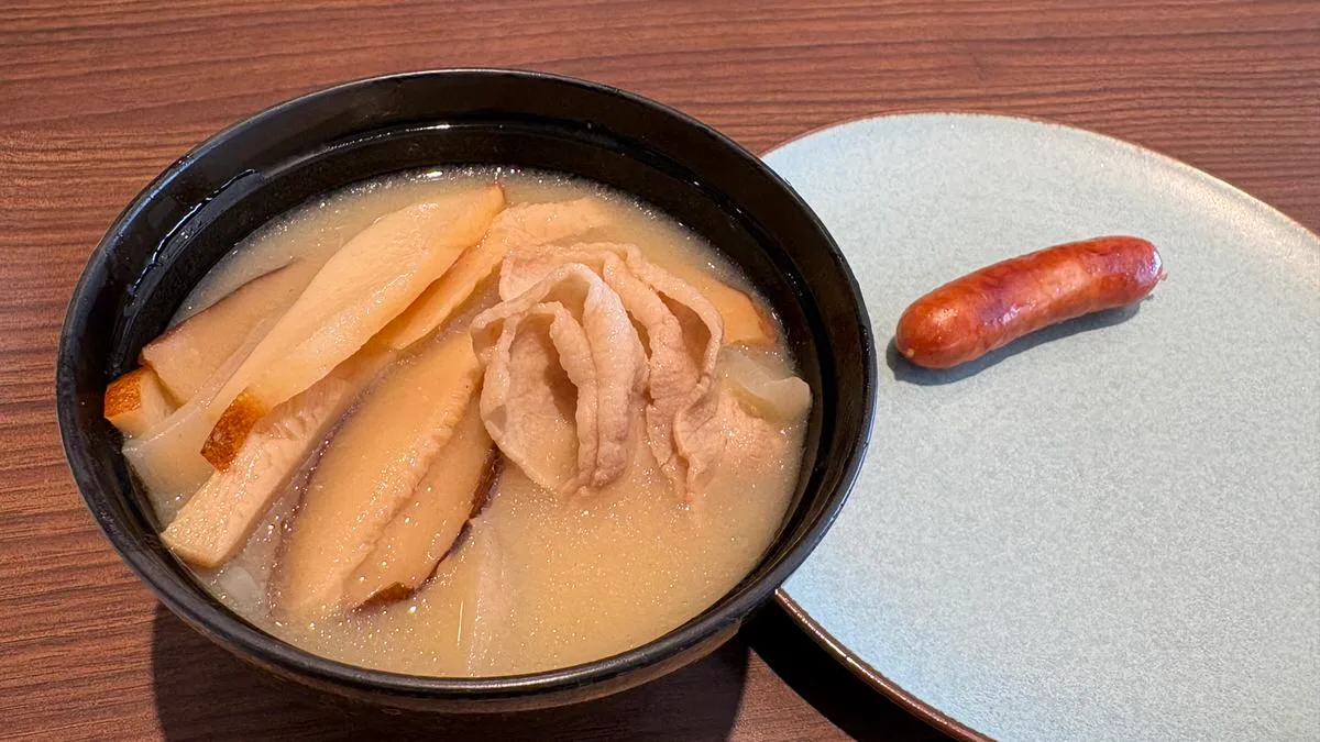A bowl of inamuduchi, a pale miso soup with sliced mushrooms and a folded slice of meat, beside a single browned sausage on a grey plate
