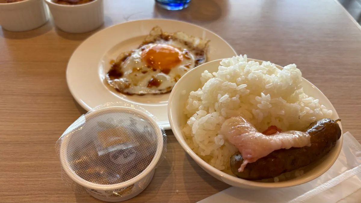 A fried egg with soy sauce on a plate, a bowl of rice topped with bacon and a browned sausage, and a lidded cup of natto